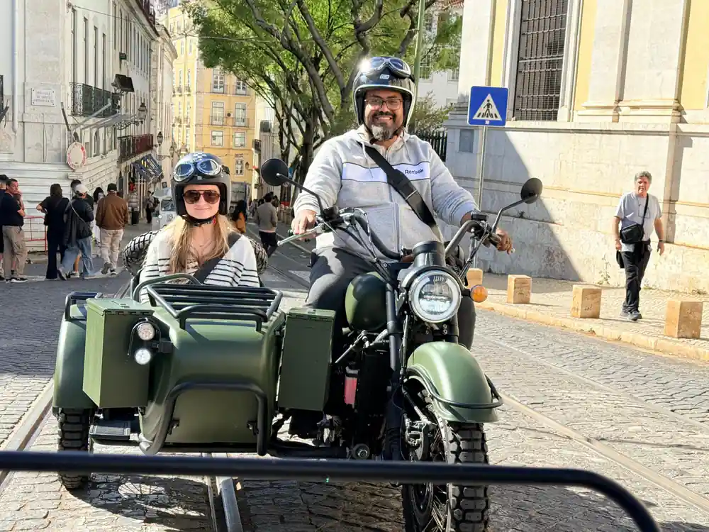 A couple riding an olive green sidecar up a street shaded by trees