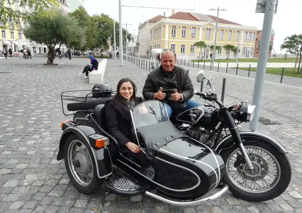 A couple seated in a black sidecar on a cobbled street below a yellow building