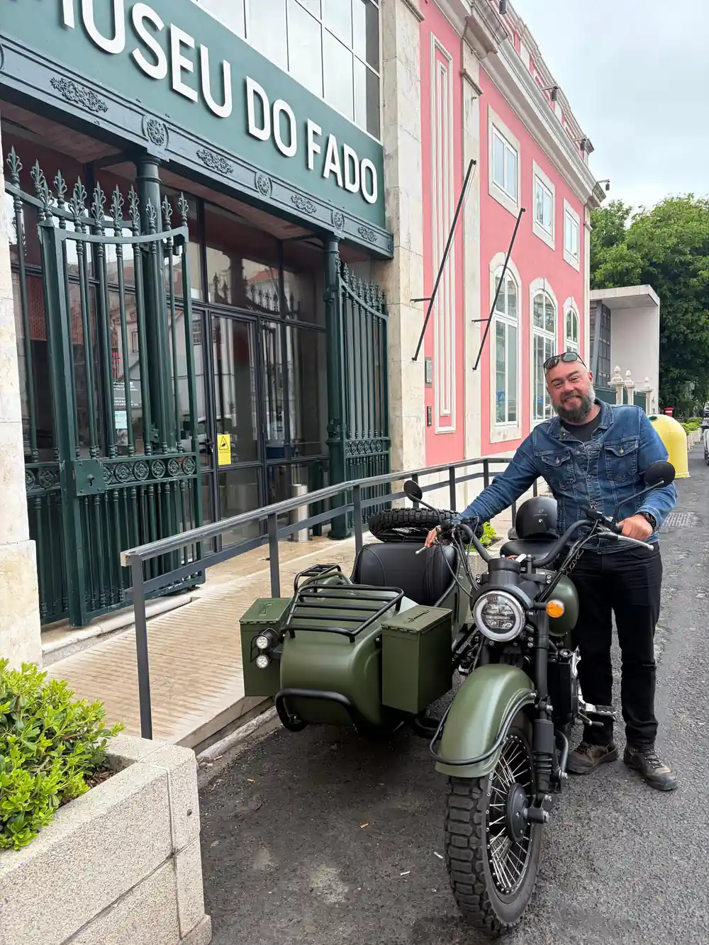 A man standing with an olive green sidecar outside a pink fado house