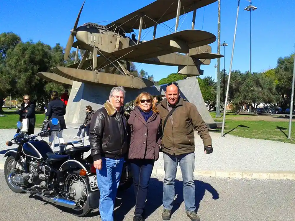 People beside a sidecar under the raised wings of the seaplane monument