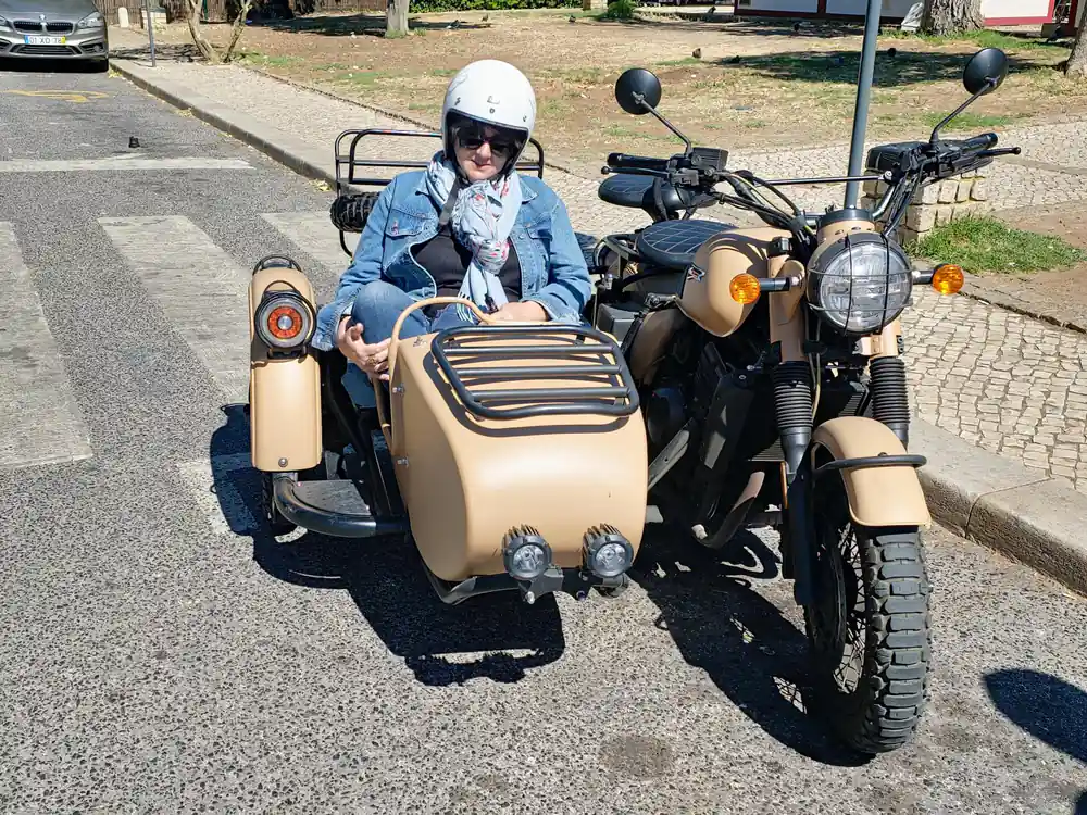 A rider seated in a tan sidecar parked in the shade of trees
