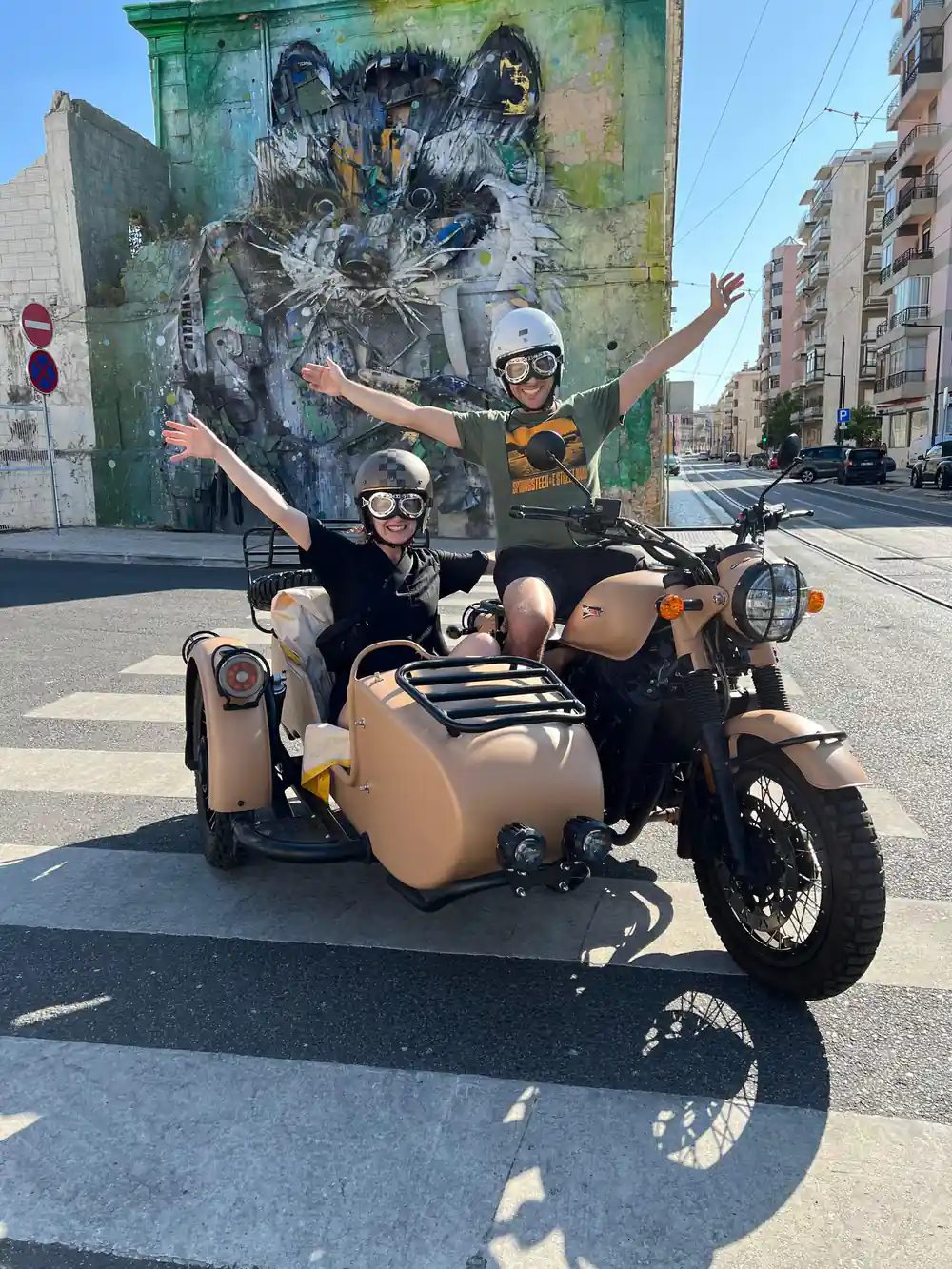 Two riders with arms raised in a tan sidecar on a street with tram rails