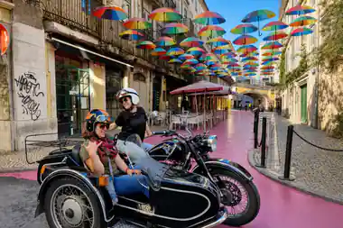Two riders in helmets in a sidecar under a street hung with coloured umbrellas