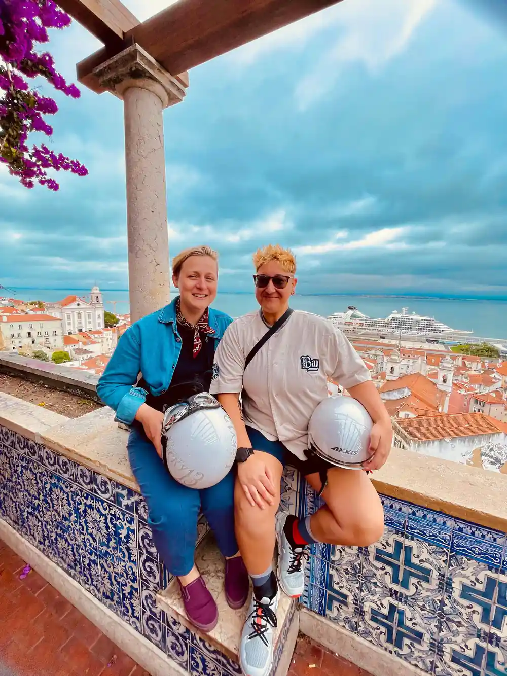 Two riders with helmets at a viewpoint above terracotta roofs and tiled walls