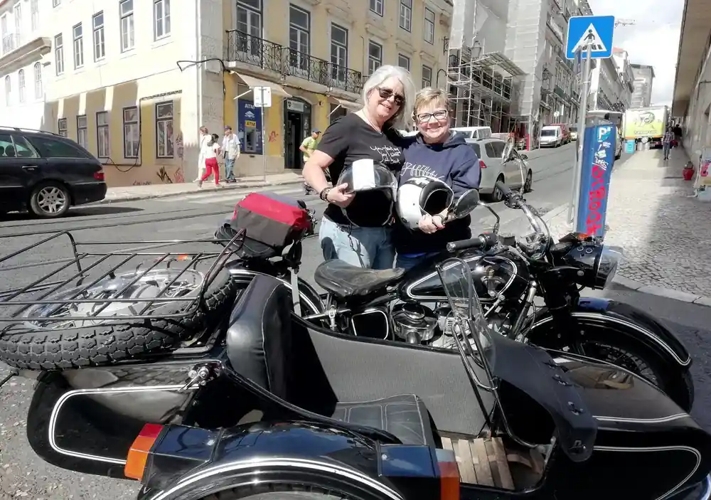 Two riders holding helmets beside a black sidecar in a city street