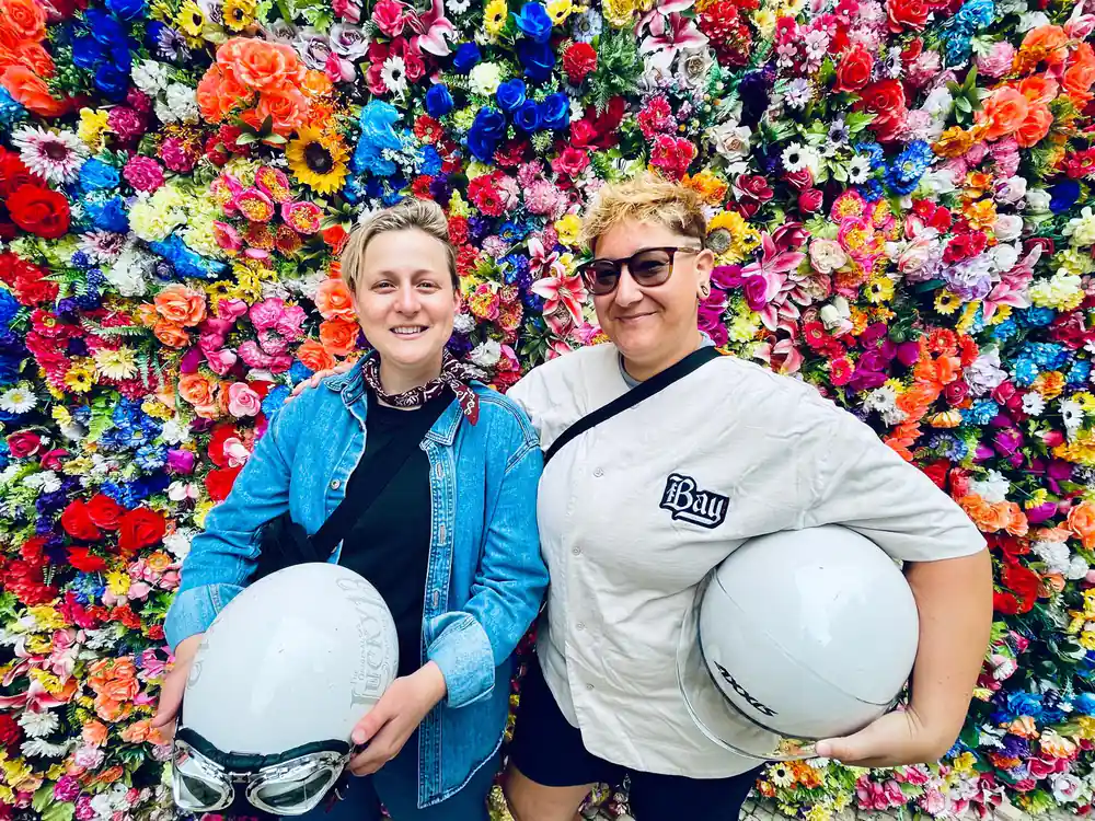 Two riders holding white helmets before a wall of artificial flowers