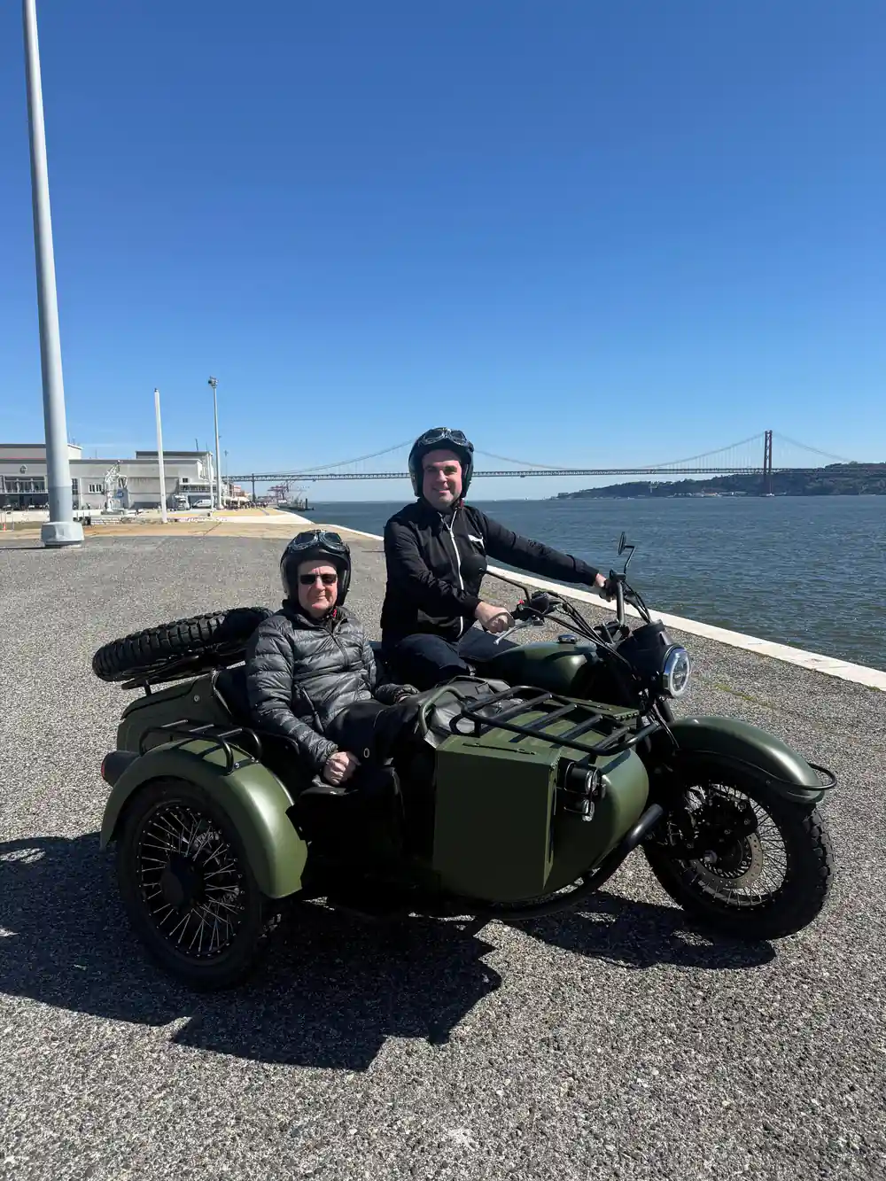 Two riders in an olive green sidecar on the riverfront beneath the bridge