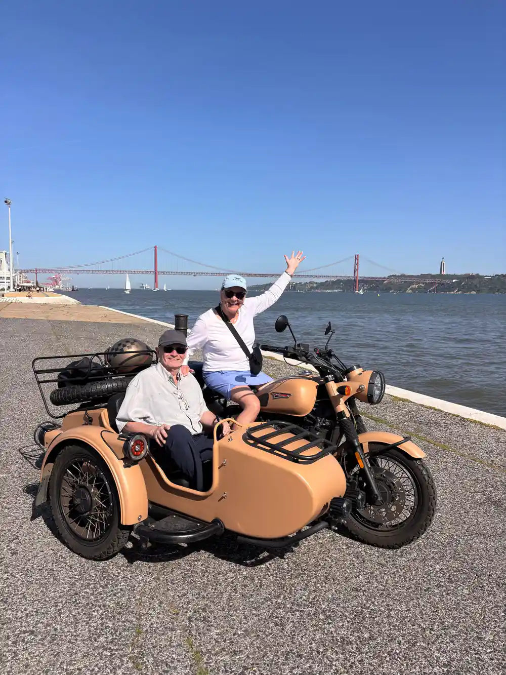Two riders waving from a tan sidecar on the riverside by the bridge