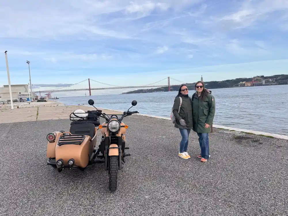 Two women beside a tan sidecar on the riverside with the bridge behind