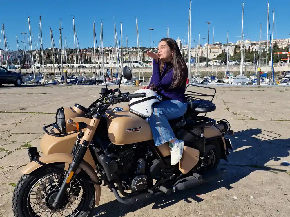 A woman seated on a tan motorcycle at the marina with masts behind