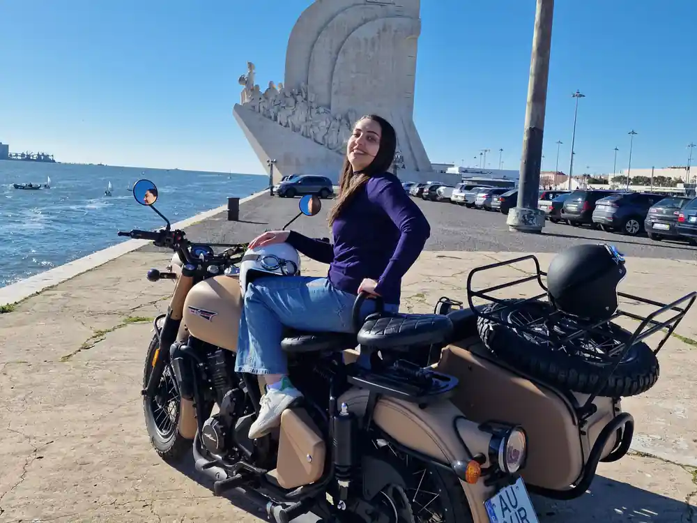 A woman seated on a tan sidecar outfit beside the Monument to the Discoveries